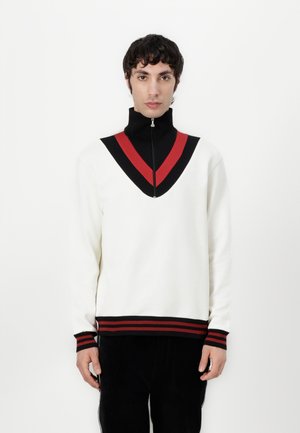 Sweatshirt blanc avec un design en V noir et rouge, col zippé et accents rayés sur les poignets et l'ourlet. Texture lisse, coupe décontractée.