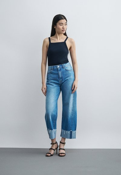 Γυναίκα με μακριά μαύρα μαλλιά που φοράει μαύρο αμάνικο τοπ, ψηλόμεσα κατεβασμένα μπλε τζιν και μαύρα πέδιλα με τακούνι, στέκεται δίπλα σε απλό τοίχο.