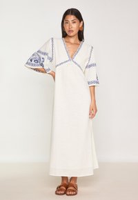 Abito maxi bianco con ricami floreali blu, scollatura a V e maniche ampie. Il materiale appare strutturato. Indossato con sandali marroni.