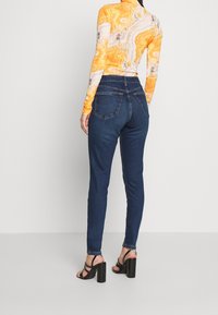 Vaqueros de mezclilla azules de corte slim, con bolsillos traseros y un ligero estiramiento. Combinados con una blusa de manga larga con estampado y sandalias negras de tacón cuadrado.