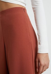 Gros plan sur une personne portant un pantalon taille haute couleur rouille et un haut à manches longues à côtes blanc, avec la main reposant sur le côté.