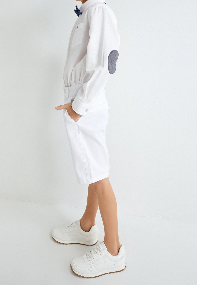 Garçon portant une chemise blanche à boutons avec un patch bleu au coude, un short blanc et des baskets blanches, debout les mains dans les poches.