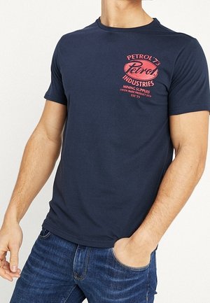 Mann trägt ein dunkelblaues T-Shirt mit rotem "Petrol Industries"-Logo und Text auf der linken Brust, kombiniert mit blauen Jeans.