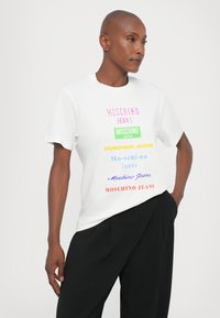 White t-shirt featuring multicolored logos and text that reads "MOSCHINO JEANS" in various fonts. Short sleeves, regular fit, smooth fabric.