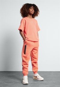 Tricou coral cu mâneci scurte, asociat cu pantaloni de trening coral cu un buzunar cu fermoar negru, purtat cu adidași albi. Textură netedă.