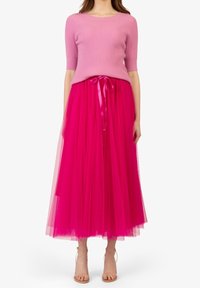 Žena v růžovém žebrovaném svetru s krátkým rukávem a jasně růžové tylové midi sukni, s béžovými sandály na kotníkovém pásku, stojící na bílém pozadí.