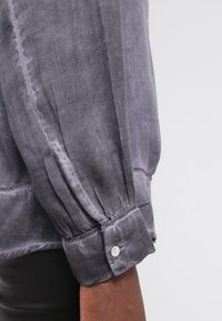 Blusa in seta grigia con maniche arricciate, polsini con bottone e texture liscia. Presenta una leggera lucentezza e un sottile dettaglio di cucitura.