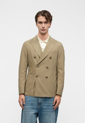 Junger Mann, der ein beiges, zweireihiges Blazer über einem weißen Poloshirt und hellblauen Jeans trägt, steht vor einem schlichten weißen Hintergrund.