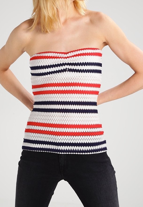 Femme portant un haut tricoté bustier avec des rayures horizontales rouges, blanches et bleu marine et un pantalon noir, debout les mains sur les hanches.