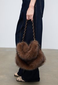 Brun taske i imiteret pels med kæderem, der har et rundt design og blød tekstur, båret af en person iført mørke denimbukser.