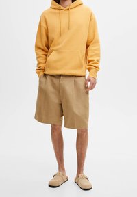 Sudadera amarilla con capucha y un bolsillo frontal; combinada con pantalones cortos de pinzas beige y zapatos de slip-on color crema. Diseño simple y corte relajado.