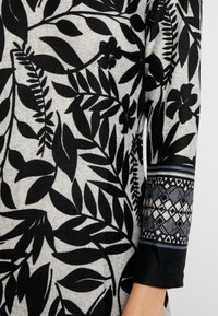 Motif floral et de feuilles noires sur un fond gris, avec un poignet noir texturé orné d'accents géométriques. Conception à manches longues.
