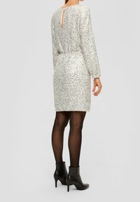 Silverfärgad paljettklänning med långa ärmar, figursydd midja och en nyckelhålskärning bak, i kombination med svarta ankelskor och genomskinliga strumpbyxor.