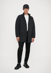 Giovane uomo che indossa un giubbotto imbottito nero, un cappellino nero, pantaloni neri e sneakers nere in piedi contro uno sfondo bianco.