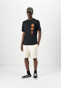 Un tricou negru cu grafică portocalie, pantaloni scurți texturati de culoare crem și pantofi sport negri. Șapca este de tip camuflaj cu un design structurat.