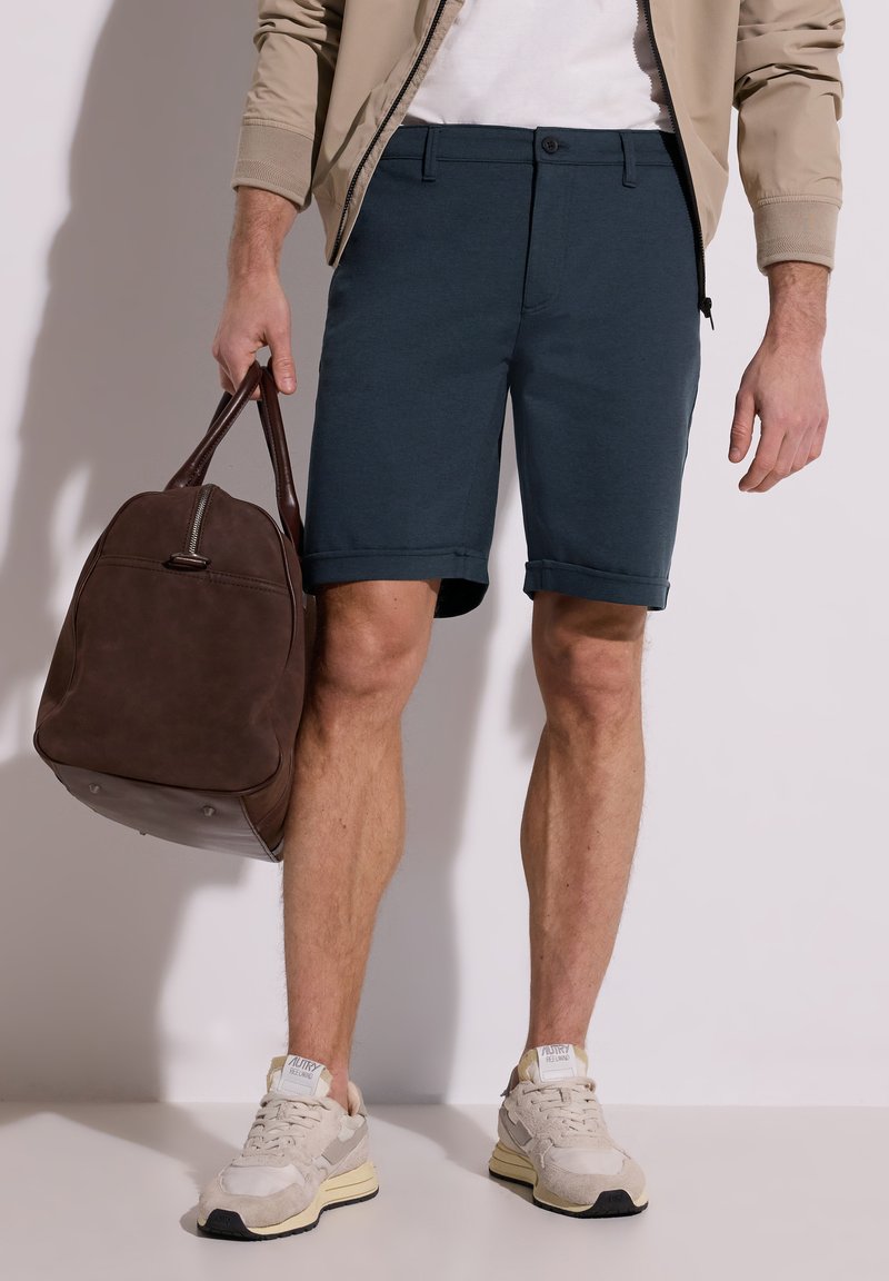 Homme portant un short bleu marine, une veste beige, des baskets blanches, tenant à ses côtés un sac en cuir marron foncé.