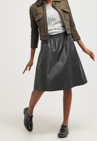 Persona vestida con una chaqueta corta verde oliva, camiseta gráfica gris, falda de cuero negra hasta la rodilla y zapatos negros brillantes con cordones, posando con los brazos extendidos.