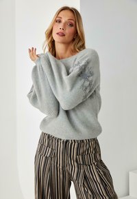 Suéter de punto gris claro, de corte holgado y con un patrón texturizado. Presenta un sutil bordado floral en una manga. Combinado con pantalones a rayas.