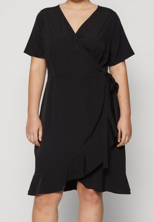 Femme portant une robe portefeuille noire à manches courtes, col en V, ceinture sur le côté et ourlet volanté, debout devant un fond clair uni.