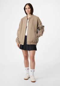 Beige übergroße Bomberjacke mit gerippten Bündchen und Saum, versehen mit einem Logo-Patch. Getragen über einem cremefarbenen Oberteil, kombiniert mit einem plissierten schwarzen Rock.