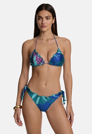 Donna che indossa un bikini blu e viola con volant e slip con lacci ai lati, con un braccialetto d'oro al polso sinistro, in piedi davanti a uno sfondo semplice.