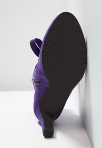 Botín de ante púrpura con un tacón alto, que presenta una cremallera lateral y una suela de goma negra texturizada con el nombre de la marca "Tamaris" en relieve.