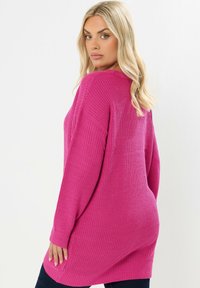 Przytulny, oversizowy różowy sweter wykonany z dzianiny, z prążkowaną teksturą, opadającymi ramionami oraz długimi rękawami. Charakteryzuje się luźnym krojem i bocznymi rozcięciami na dolnym brzegu.