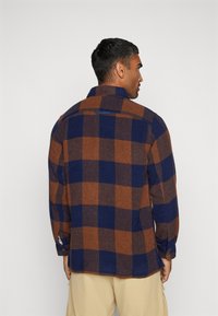 Cămașă din flanel cu carouri maro și bleumarin, cu guler, mâneci lungi și nasturi la mansete. Textură moale, cu o croială lejeră și fante laterale.