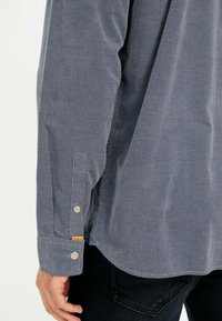 Langarmhemd aus dunkelgrauem Cord mit strukturiertem Muster. Verfügt über Knopfmanschetten und ein kleines Etikett-Detail nahe dem Saum.