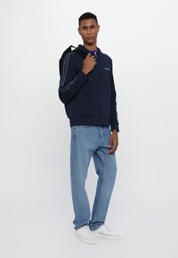 Felpa blu navy con strisce bianche, jeans a vestibilità rilassata in azzurro chiaro e sneakers bianche. Il modello tiene uno zaino nero.