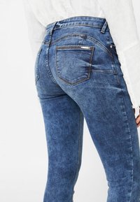 Vaqueros de mezclilla azul con un diseño ajustado, costuras detalladas y dos bolsillos traseros. La tela tiene una textura ligeramente desvaída con un acabado suave.