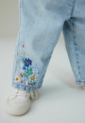 Lyseblå denimbukser med farverig blomsterbroderi ved kanten. Båret med hvide sneakers, hvilket fremhæver en afslappet og casual pasform.