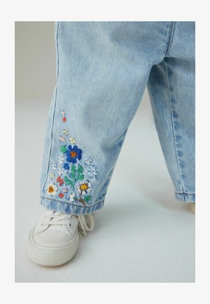 Jeans din denim albastru deschis cu broderie florală colorată la tiv. Purtate cu adidași albi, evidențiind un fit casual, relaxat.