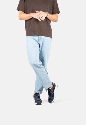 Personne se tenant debout avec les jambes croisées, portant un jean bleu clair, des baskets sombres, un t-shirt marron, et une montre sur un fond blanc.