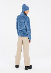 Giacca in pile blu e morbida con colletto alto, abbinata a pantaloni beige a gamba larga e scarpe chunky nere; berretto di maglia blu in testa.