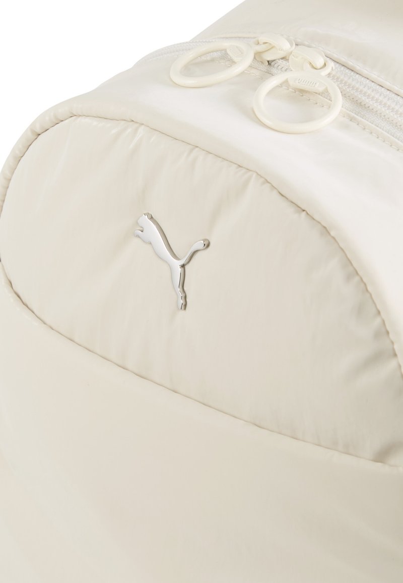 Mochila de color crema claro, fabricada en nailon suave, con un destacado logo plateado de Puma y dos tiradores de cremallera con acentos circulares.