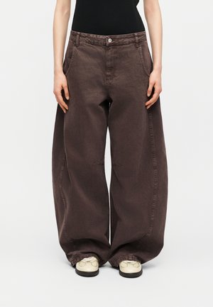 Άτομο που φοράει υπερμεγέθη, σκούρο καφέ παντελόνια με φαρδιά πόδια και πλαϊνές τσέπες, συνοδευόμενα από εκρού αθλητικά παπούτσια, στέκεται μπροστά σε ουδέτερο φόντο.