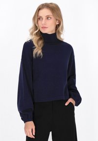 Granatowy sweter z golfem, z długimi, balonowymi rękawami i prążkowanym kołnierzem. Miękka tekstura, krótkie wykończenie i luźny krój. W zestawie z czarnymi spodniami.