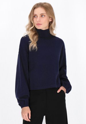 Maglione a collo alto di colore blu navy con maniche lunghe a palloncino e colletto a costine. Texture morbida, design corto e vestibilità rilassata. Abbinato a pantaloni neri.