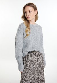Hellgrauer Strickpullover mit langen Ärmeln, der eine lockere Passform hat. Kombiniert mit einem schwarzen Blumenrock und einem schwarzen Gürtel mit einer goldenen Schnalle.