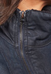 Jeansjacka med metalldragkedja och "RAW"-etikett, mörkblått tyg och en mjuk krage med textur.