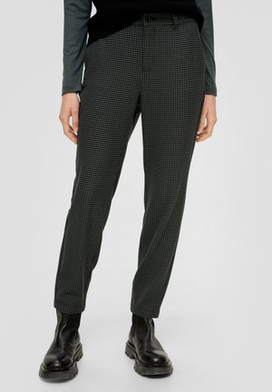 Frau trägt schwarze und graue Slim-Fit-Hahnentritt-Hose, schwarze klobige Stiefel und dunkles langärmeliges Oberteil, steht vor einem einfarbigen Hintergrund.