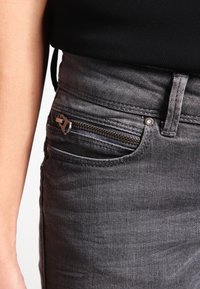 Los jeans de mezclilla gris cuentan con un bolsillo con cremallera, detalles de costura sutiles y una textura suave. La tela tiene un ligero brillo.