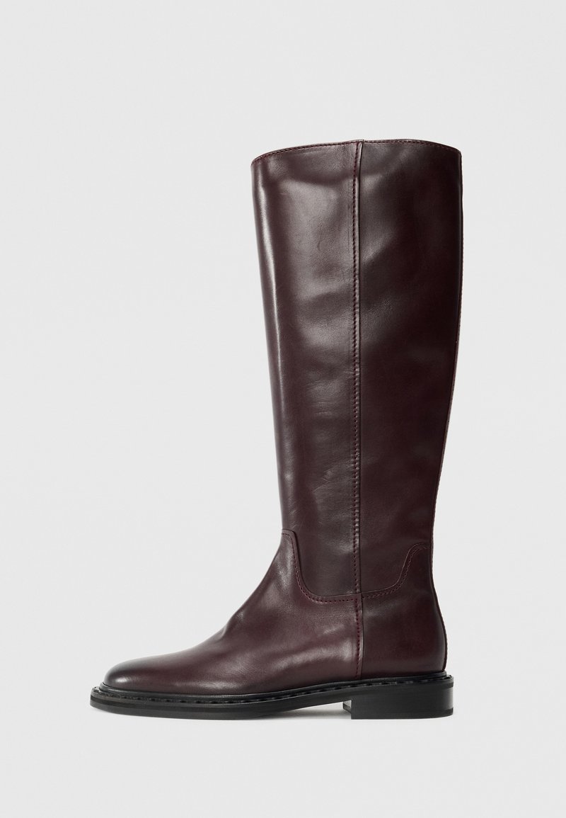 Botte en cuir bordeaux hauteur genou avec une texture lisse, bout rond et des détails de couture minimaux. Elle dispose d'une semelle noire plate.