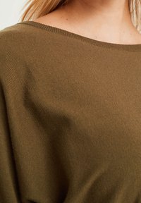 Pull en vert olive en tricot côtelé avec un large col bateau. Texture lisse avec des variations de couleur subtiles.