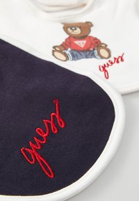 Cotton bib set featuring a cream-colored bib with a brown bear graphic, and a dark navy bib with red embroidered "guess" text. White trim accents.