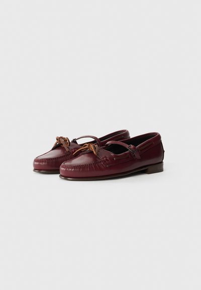 Mocasines de cuero en burdeos oscuro, con punta redonda, detalle de cordones con dos lazos de cuero marrón y suela plana con un tacón mínimo.