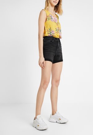Femme portant un haut sans manches jaune à motifs floraux, un short en jean noir taille haute et des baskets blanches, debout devant un fond blanc.