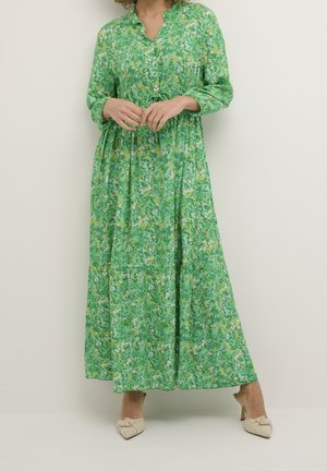 Woman wearing a long green floral dress with buttons down the front, standing with hands clasped and beige heeled mules.