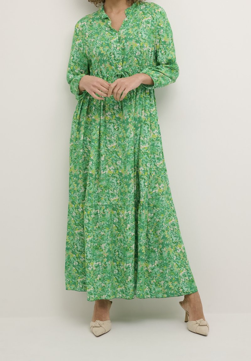 Femme portant une longue robe verte à fleurs avec des boutons sur le devant, debout les mains jointes, chaussée de mules à talons beiges.